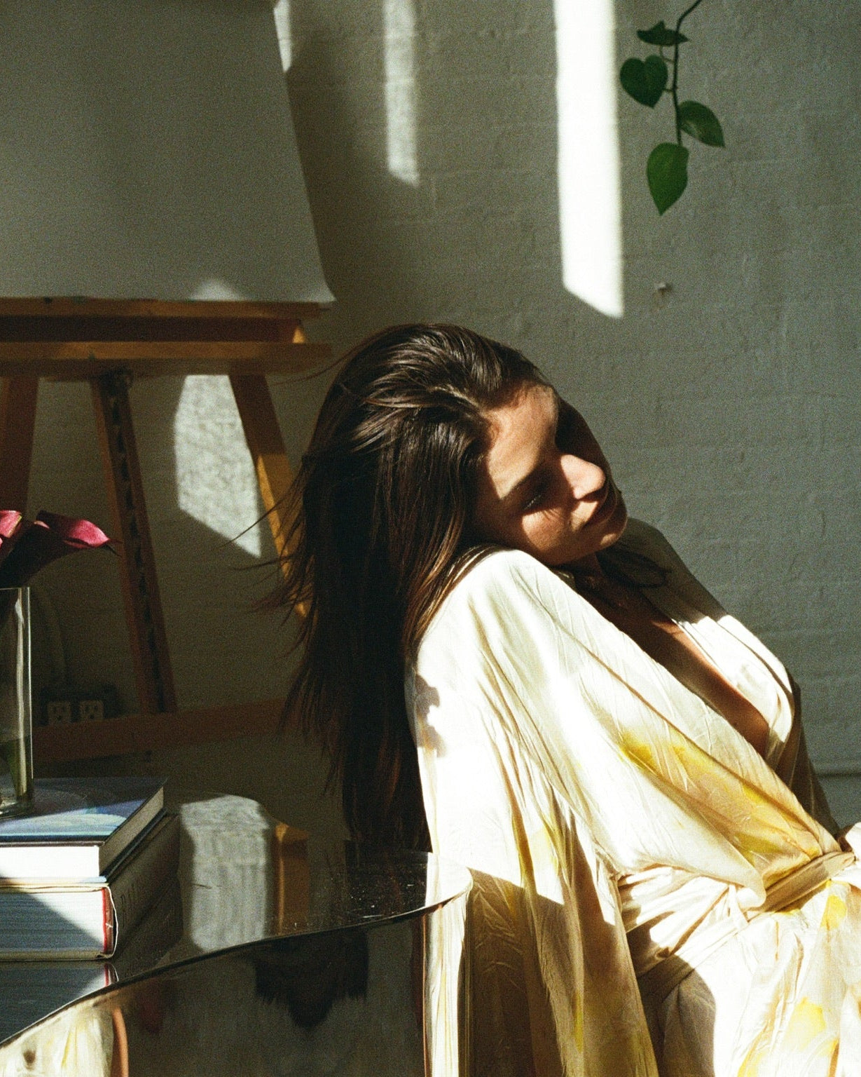 Model n a white robe sitting in a sunlit room with plants and books.